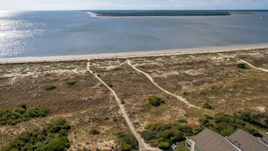 Vista aérea de una zona costera con una playa arenosa, una pequeña isla a lo lejos y un camino pavimentado que conduce a un edificio.