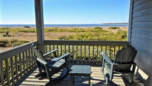 A covered deck with Adirondack chairs and a small table, overlooking a coastal landscape. - Seabrook Exclusives | 1377 Pelican Watch (Johns Island)