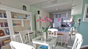 Une cuisine moderne avec des meubles blancs, un coin repas doté d’une table en verre et de chaises blanches, et une bibliothèque remplie de livres et d’objets décoratifs.