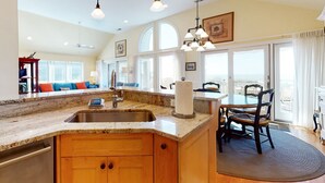 Una cocina amplia con encimeras de granito, armarios de madera y una isla grande. Hay un comedor con una mesa y sillas, y vistas al exterior a través de grandes ventanales.