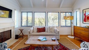 Una sala de estar con un sofá blanco, una mesa de centro de madera, una chimenea y ventanas amplias con persianas.