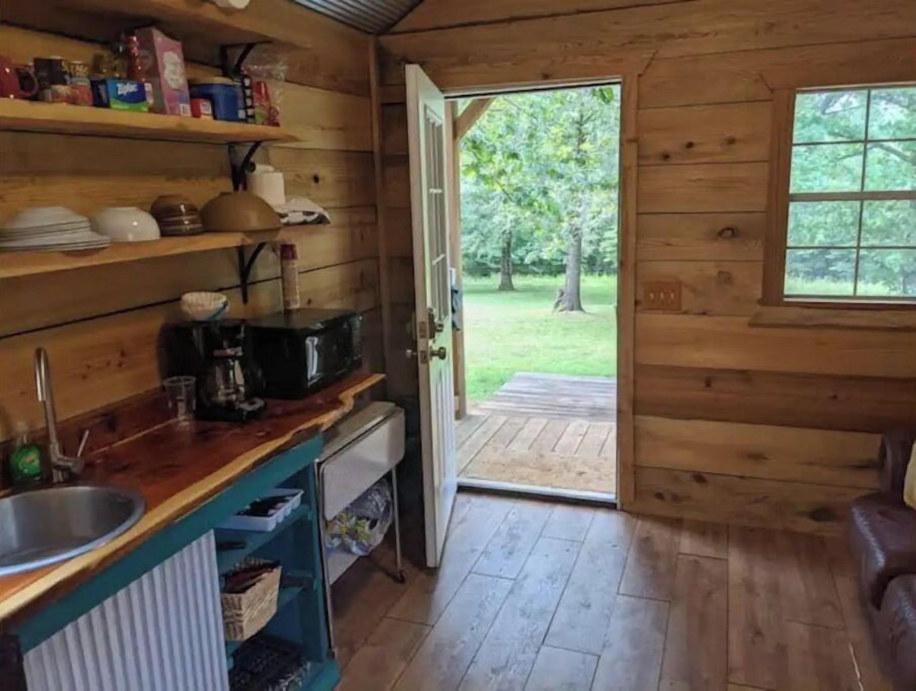 Eine Holzhütte mit Küchenbereich, der Regale mit Geschirr und eine Kaffeemaschine sowie ein Spülbecken beinhaltet, mit Blick auf eine grüne Wiese durch eine offene Tür.