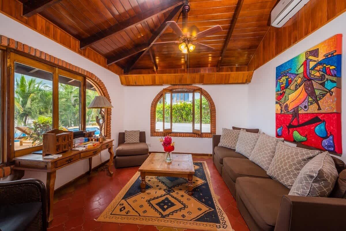 Una sala de estar acogedora con techo de madera, ventilador de techo, un ventanal grande, un cuadro abstracto colorido, un sofá, una mesa de centro y un escritorio con lámpara.