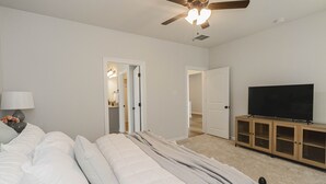 A bedroom with a bed, a ceiling fan, a TV on a wooden cabinet, and a door leading to another room. - Cozy NEW Home in the Heart of Lafayette! (Lafayette)