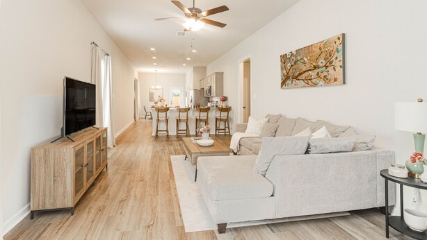 A spacious living room with a sofa, a coffee table, and a TV on a wooden cabinet. There is a ceiling fan, a painting on the wall, and a kitchen in the background. - Cozy NEW Home in the Heart of Lafayette! (Lafayette)
