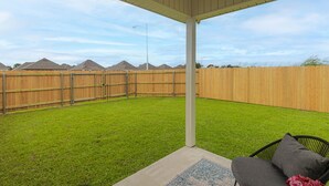 A covered patio area with a green lawn, a wooden fence, and a single outdoor chair. - Cozy NEW Home in the Heart of Lafayette! (Lafayette)