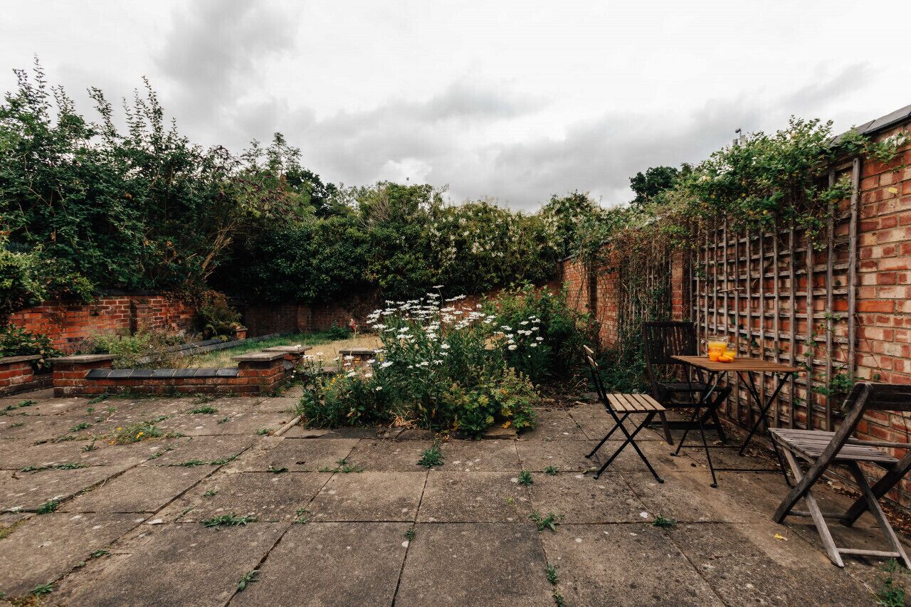 Un patio con pared de ladrillo, una mesa de madera con sillas y un área jardín con flores blancas y follaje.