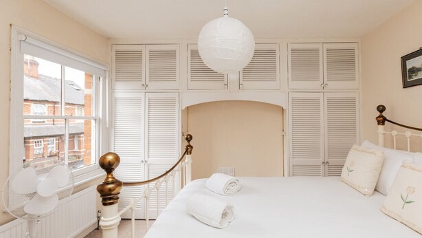 A bedroom with a bed, white shutters, a radiator, a hanging lamp, and a window with a view of buildings.