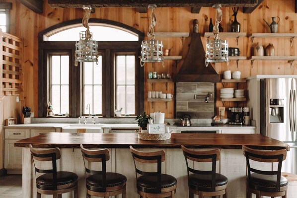 A rustic kitchen with wooden walls, a large island with bar stools, hanging light fixtures, and various kitchen items on shelves.