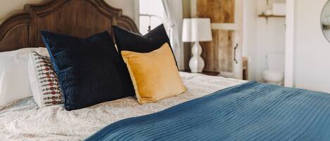 A bedroom with a wooden headboard, a bed with patterned pillows, a blue blanket, and a wooden door with a white curtain.