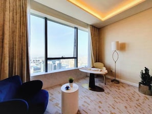 Une chambre d’hôtel moderne avec une grande fenêtre, un canapé bleu, une petite table ronde, un lampadaire et une plante en pot.