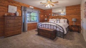 A bedroom with a wooden floor, a bed with a metal frame, a dresser, a window with curtains, a ceiling fan, and a mirror on the wall. - Cabin I at Safe Harbor Brady Mountain on Lake Ouachita! (Royal)