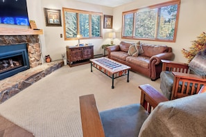 A cozy living room with a fireplace, leather sofa, wooden coffee table, and large windows with blinds. - Spacious Breck Hideaway. Hot Tub. On Town Bus. (Breckenridge)