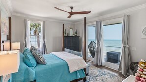 Ein Schlafzimmer mit Bett, Deckenventilator, Nachttisch, Lampe, Fenster mit Vorhängen, Blick auf das Meer und einer Tür, die nach draußen führt.