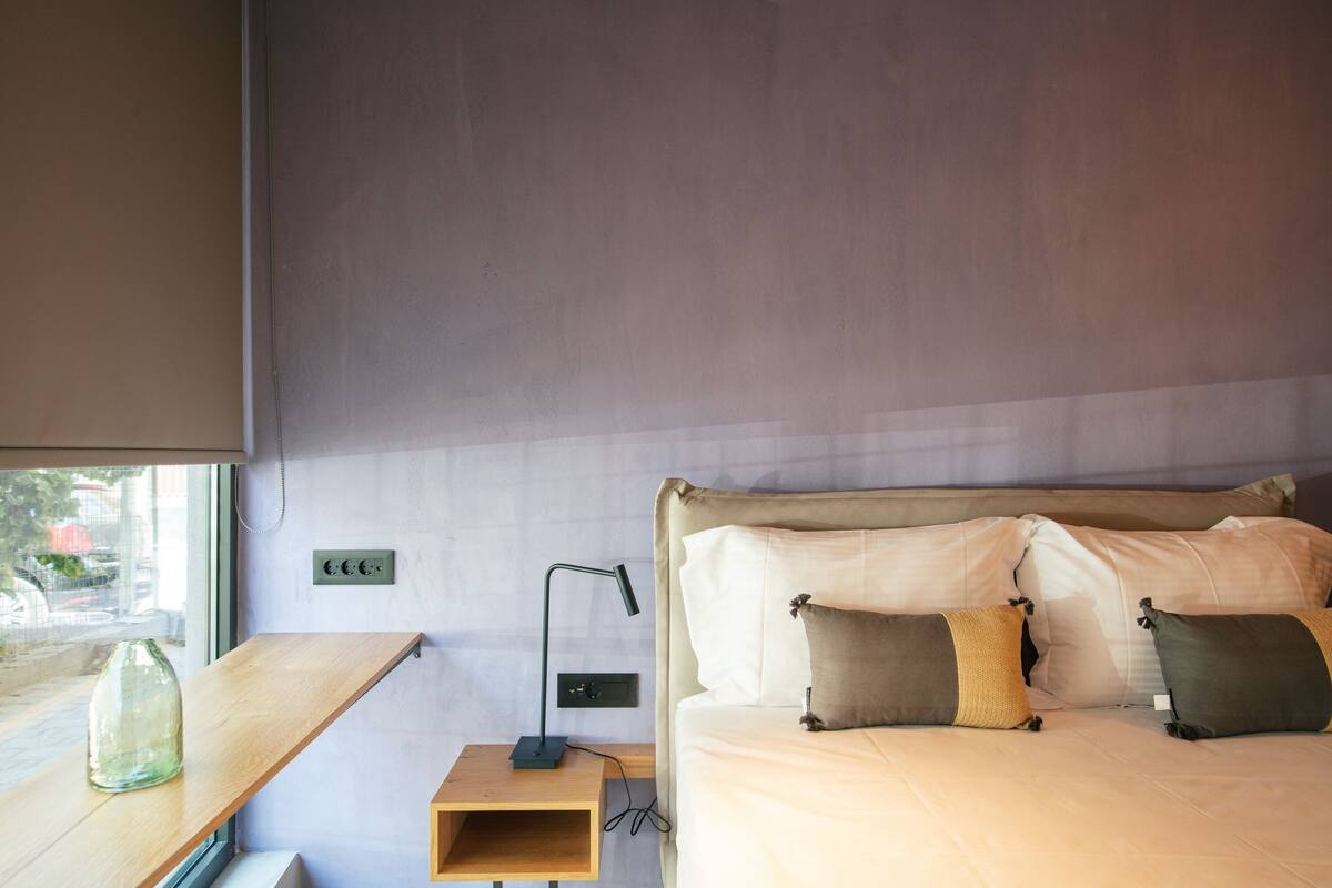 Un dormitorio con cama, almohadas, una mesita de noche y una lámpara, además de una ventana.