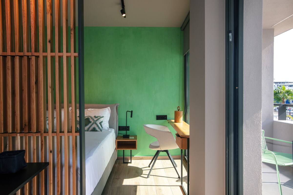 Un dormitorio con pared verde, un separador de madera, cama con ropa blanca, un escritorio con silla y ventana con vista al exterior.