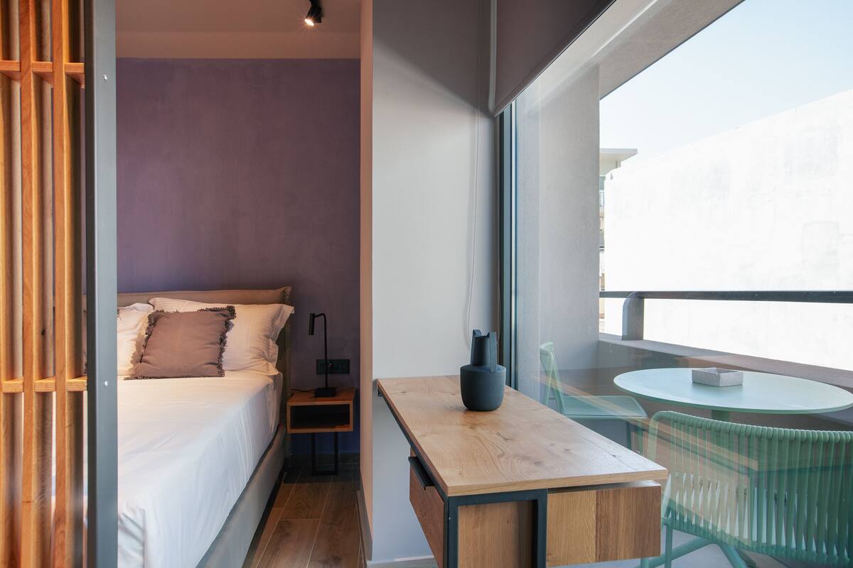 Habitación pequeña con cama, una mesita, una silla y ventana con vistas a edificios.