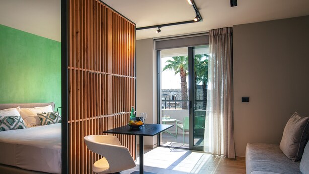 A modern hotel room with a bed, a small table with a bottle and glasses, a chair, a sofa, and a balcony with a view of palm trees. - Souda Plaza Collection (Chania)