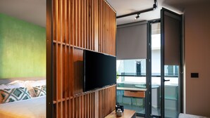 A modern bedroom with a bed, a wooden panel wall, a TV, a balcony with a glass door, and a small table with a bottle. - Souda Plaza Collection (Chania)