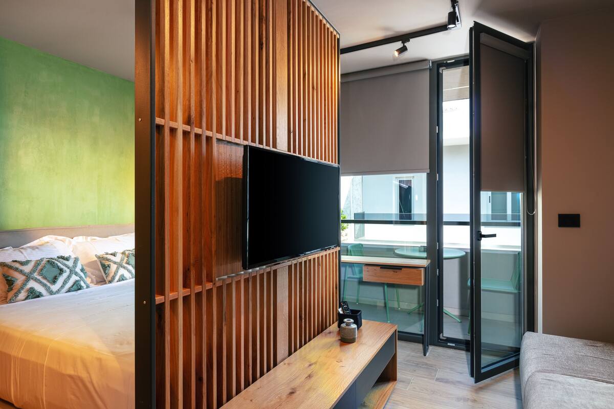 Un dormitorio moderno con una cama, un panel de pared de madera, un televisor, un balcón con puerta de cristal y una pequeña mesa con una botella.