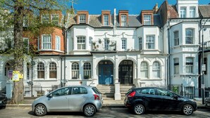 A row of white and red brick townhouses with a central blue door, parked cars, and a tree on the left. - JOIVY Captivating 1-Bed Flat In Fulham (London)