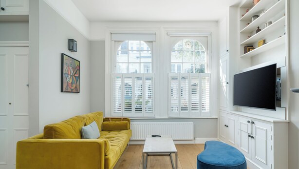 A living room with a yellow sofa, a blue ottoman, a white coffee table, and a TV on a white cabinet. - JOIVY Captivating 1-Bed Flat In Fulham (London)