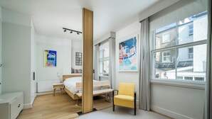 A modern bedroom with a wooden bed, a yellow chair, a large window, and a radiator. - JOIVY Captivating 1-Bed Flat In Fulham (London)