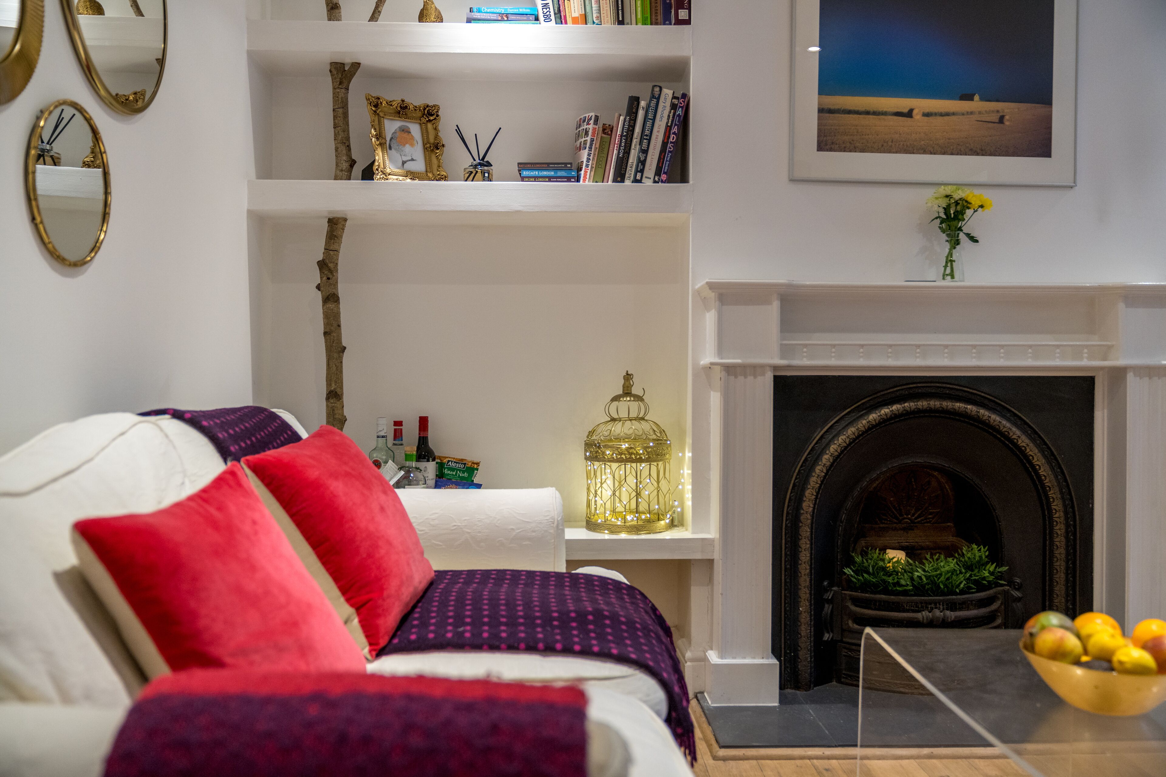 A living room with a white sofa, red and purple cushions, a fireplace, a bookshelf, and a TV on the wall.
