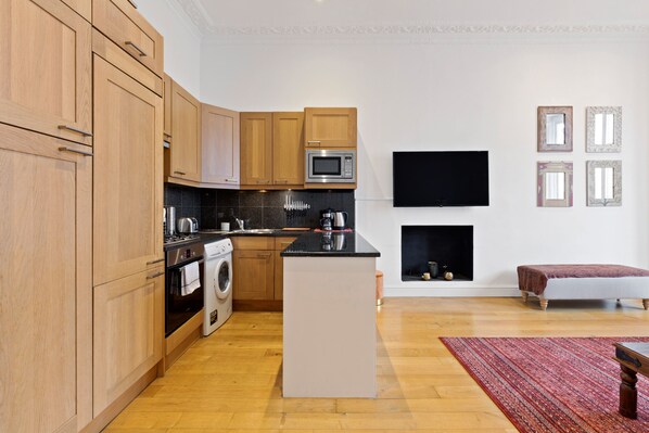 A modern kitchen with wooden cabinets, a microwave, and a built-in oven. There is a flat-screen TV mounted on the wall and a small seating area with a bench and a rug. - JOIVY Elegant 1-Bed Flat In Bayswater (London)