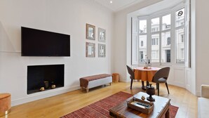 Ein modernes Wohnzimmer mit Flachbildfernseher, Kamin, Essbereich mit Tisch und Stühlen sowie großen Fenstern mit Blick auf Gebäude.