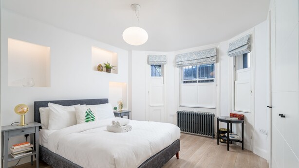 A neatly arranged bedroom with a bed, bedside tables, a radiator, and windows with curtains. - JOIVY Stylish 2-Bed Flat W/ Private Garden In Notting Hill, (London)