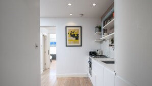 A modern kitchen with white cabinets, a stove, and a sink. There is a framed artwork on the wall and a hallway leading to another room. - JOIVY Stylish 2-Bed Flat W/ Private Garden In Notting Hill, (London)