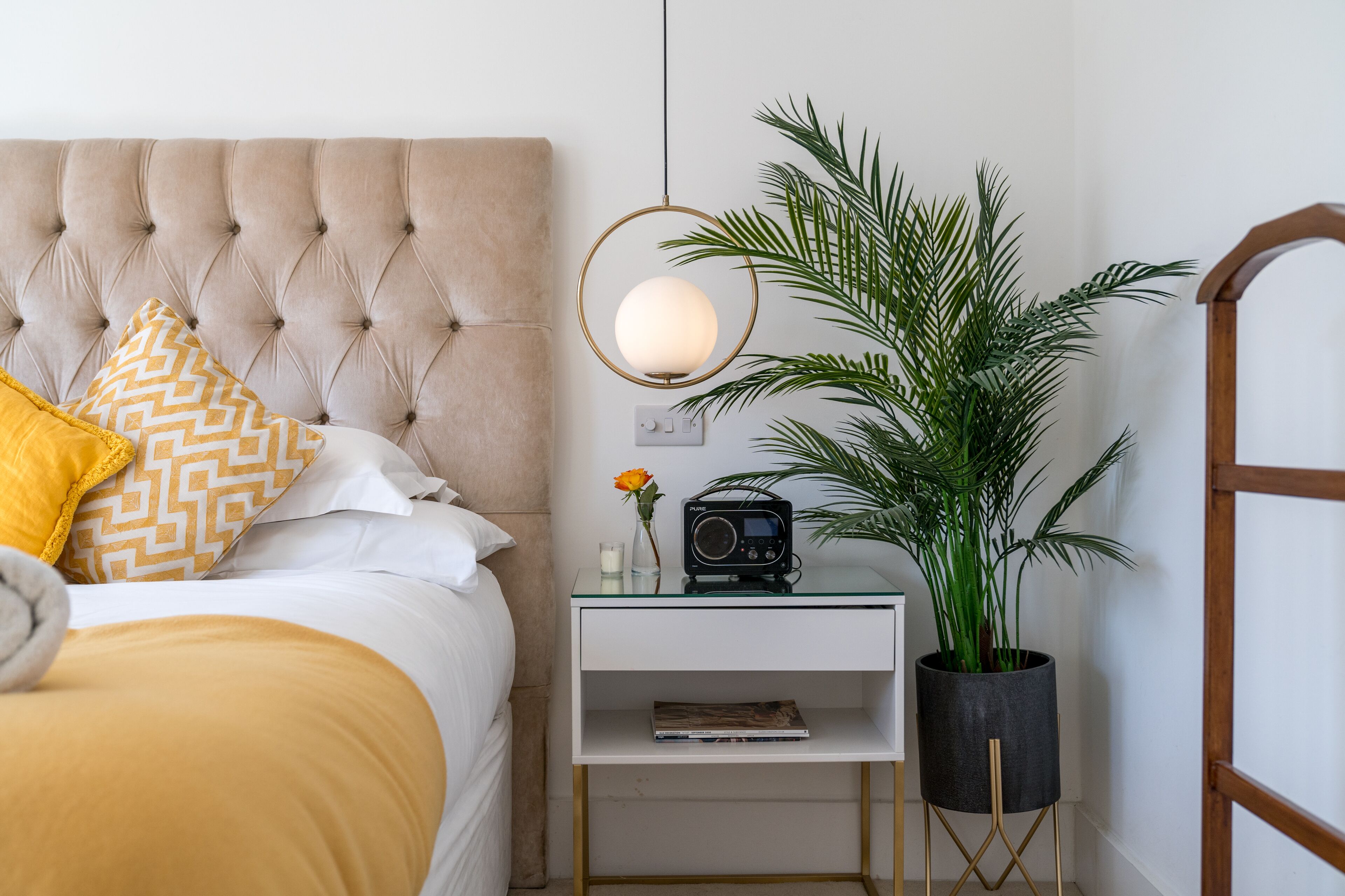 Une chambre à coucher bien rangée, avec une tête de lit capitonnée, une table de chevet dotée d’une radio, une plante en pot et une lampe de chevet.