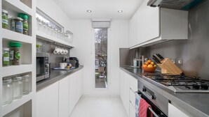 A narrow kitchen with white cabinets, a stainless steel stove, and a window overlooking a garden. - JOIVY Captivating 2-Bed Flat In Shepherd'S Bush (London)