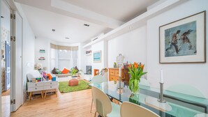 A modern living room with a glass dining table, white walls, and a large painting. - JOIVY Captivating 2-Bed Flat In Shepherd'S Bush (London)