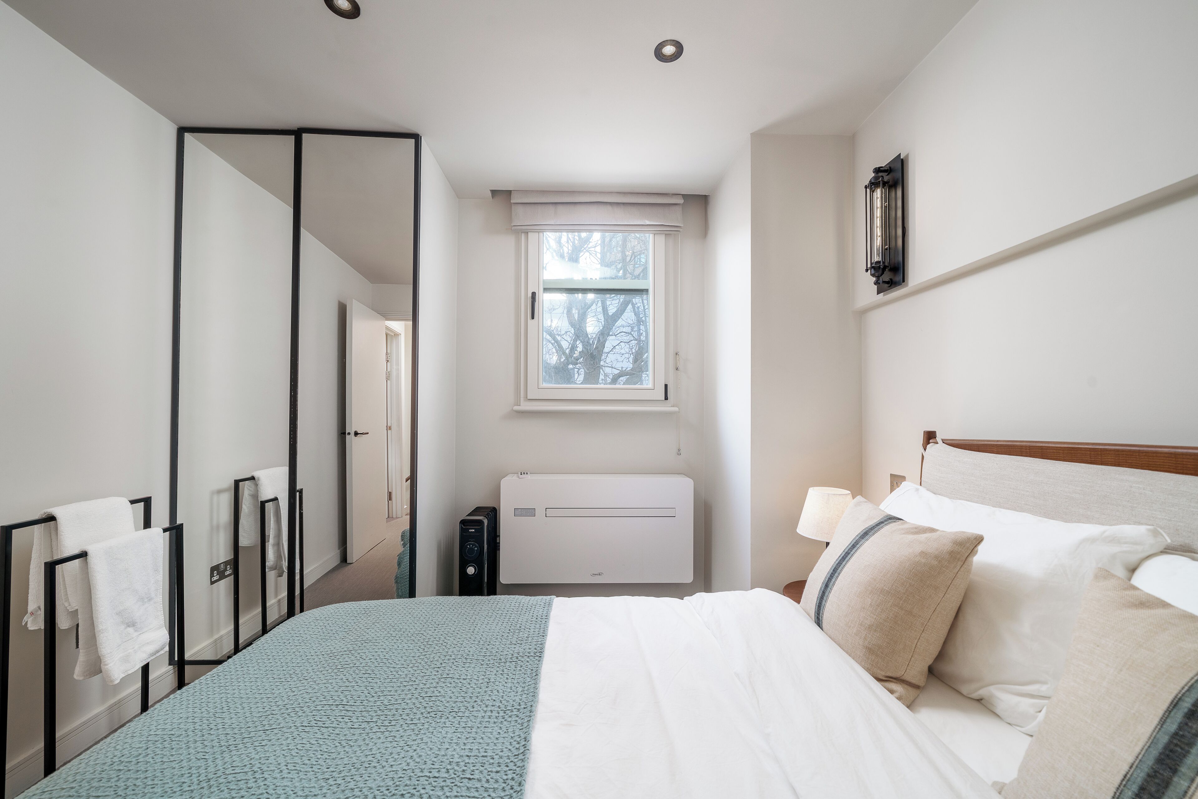 A bedroom with a bed, a bedside table with a lamp, a window, a mirror, and a radiator.