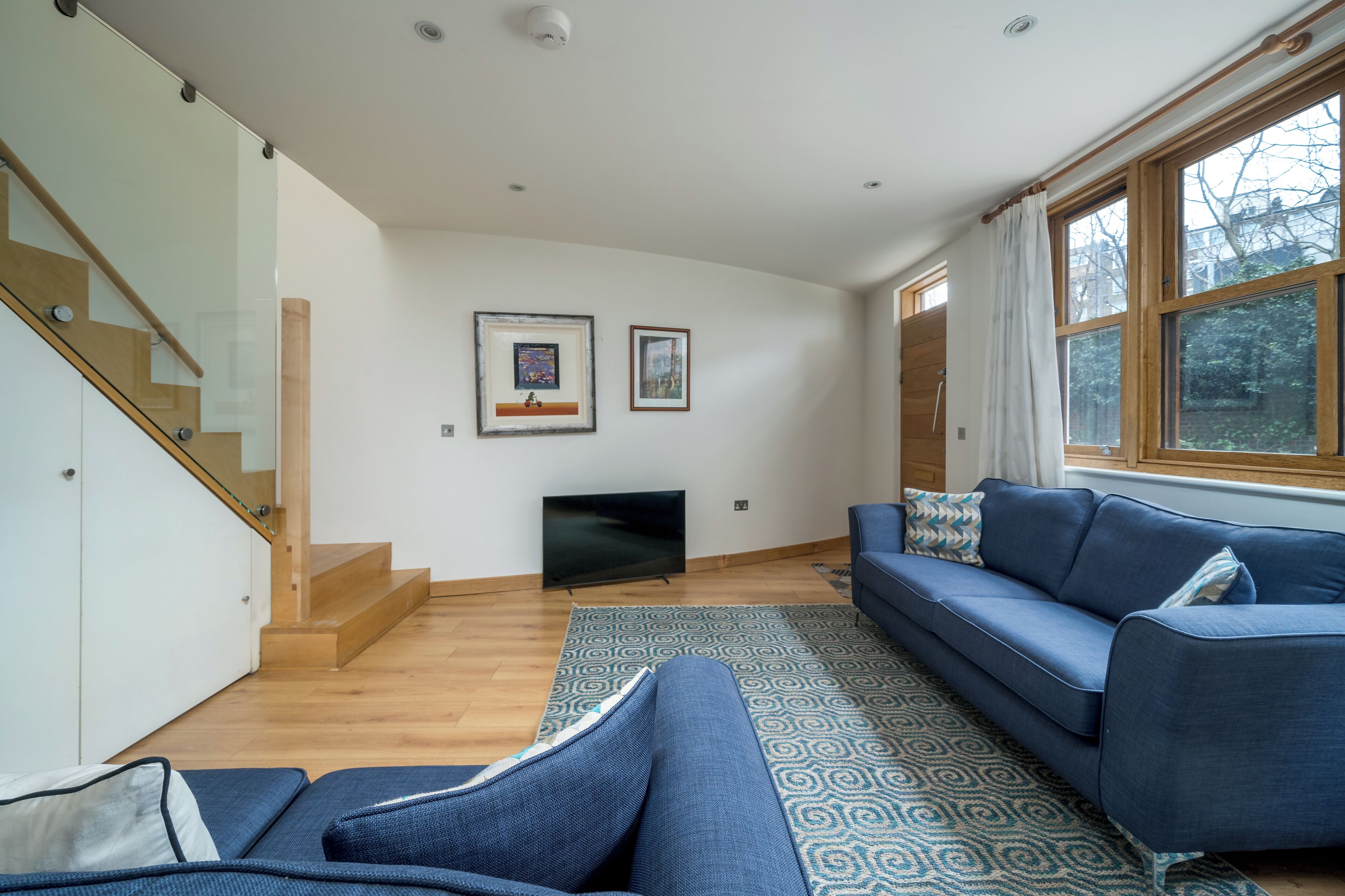 A modern living room with a blue sofa, a flat-screen TV, and a staircase with glass railings.