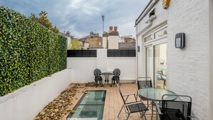 Eine kleine Terrasse mit einem Glastisch und Stühlen, einer weißen Backsteinmauer und einer Hecke.