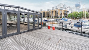 Een houten terras met glazen wanden met uitzicht op een jachthaven met boten en gebouwen op de achtergrond.