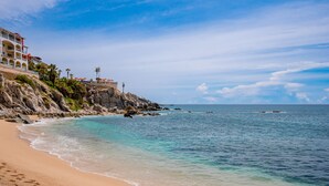 A beach with clear turquoise water, a sandy shore, and a building with balconies overlooking the ocean. - Peaceful ocean-view condo in an oceanfront community with private beach access! (Cabo San Lucas)
