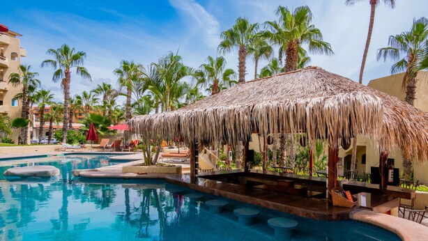 A resort pool area with a thatched-roof structure, palm trees, and lounge chairs. - Peaceful ocean-view condo in an oceanfront community with private beach access! (Cabo San Lucas)