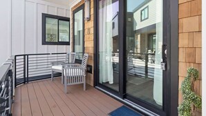 A balcony with a wooden floor, a white chair, a black railing, and a glass door leading to a building.