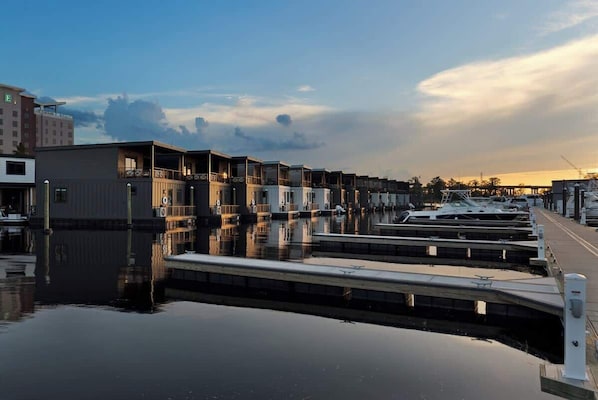 A marina with docked boats, a row of buildings with balconies, and a calm water surface reflecting the sky.