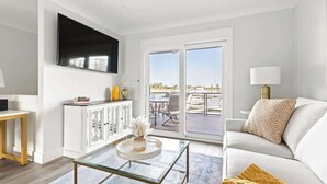 A living room with a TV, white furniture, a glass coffee table, and a view to an outdoor patio.