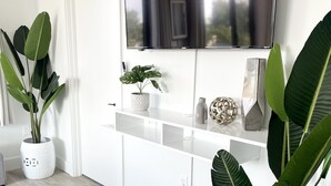 Una sala de estar moderna con un televisor de pantalla plana grande, muebles blancos y plantas verdes de gran tamaño.