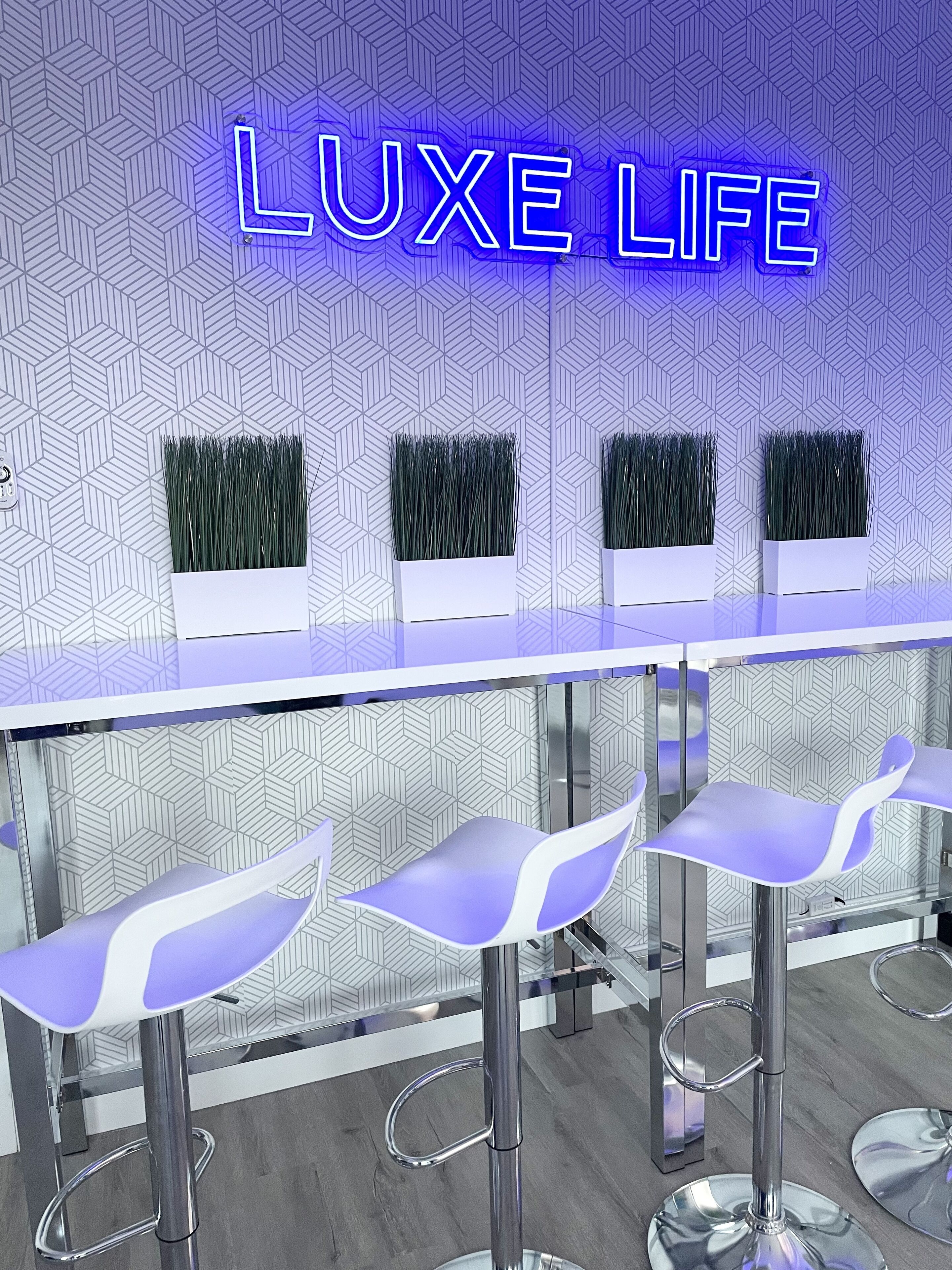 Un espace bar agrémenté d'un panneau néon sur lequel on peut lire « LUXE LIFE », de trois tabourets de bar et de jardinières décoratives.