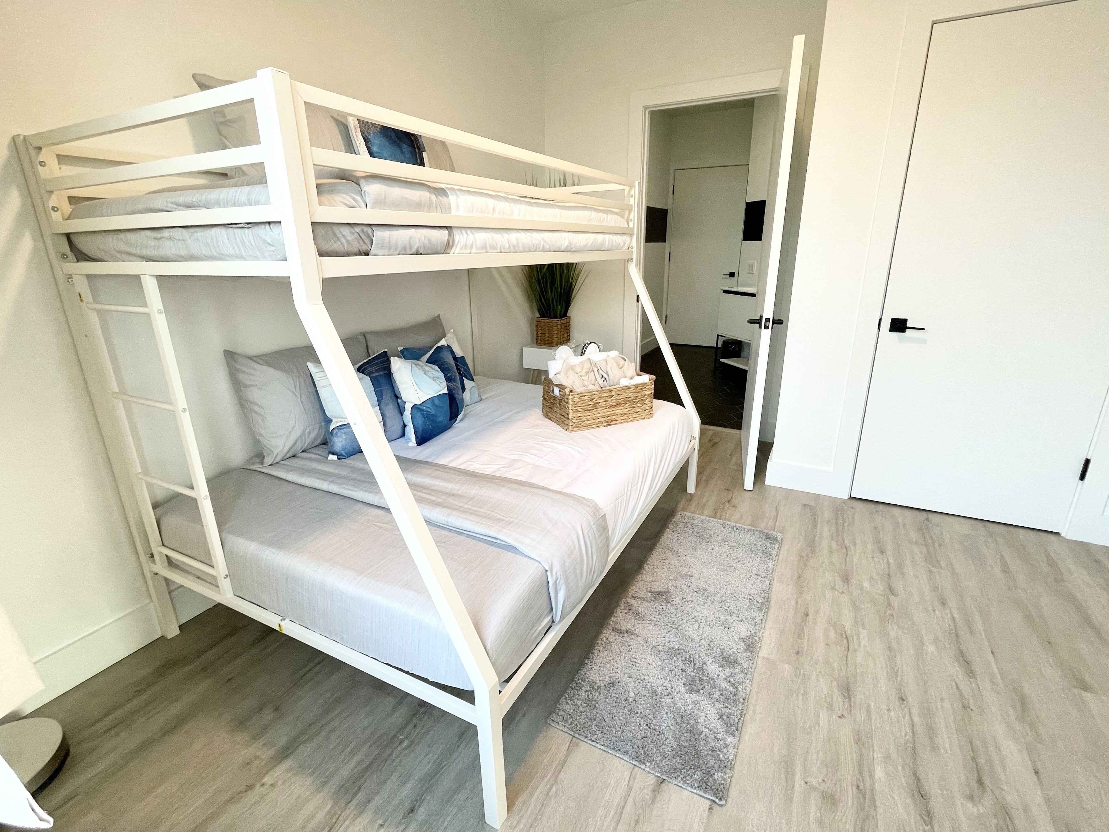 A bunk bed with a grey mattress, white frame, and blue and grey pillows. There is a grey rug on the floor and a door in the background.