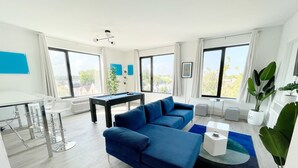 Una sala de estar moderna con un sofá azul, una mesa de billar y ventanas amplias con vistas a la ciudad.