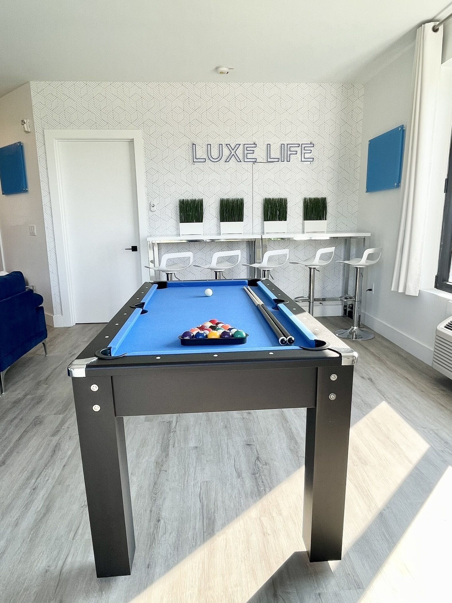 A pool table with a blue felt surface, surrounded by white chairs, in a room with a geometric patterned wall and a 'LUXE LIFE' sign.