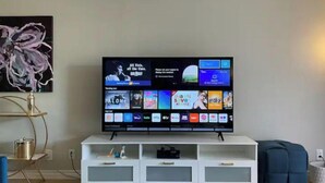 Televisão de tela plana em suporte branco com design moderno, exibindo interface de serviço de streaming com diversas opções de conteúdo.